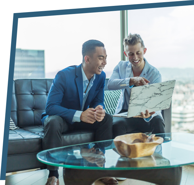 Two men in business attire are sitting on a modern sofa, smiling and discussing something on a marble-patterned laptop in a bright, glass-walled office with a city view